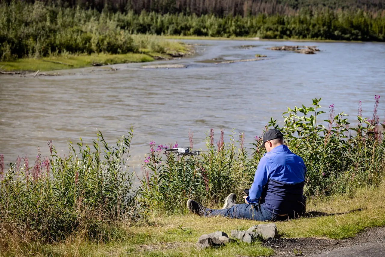 Drone operator seated by a river with a small drone hovering nearby