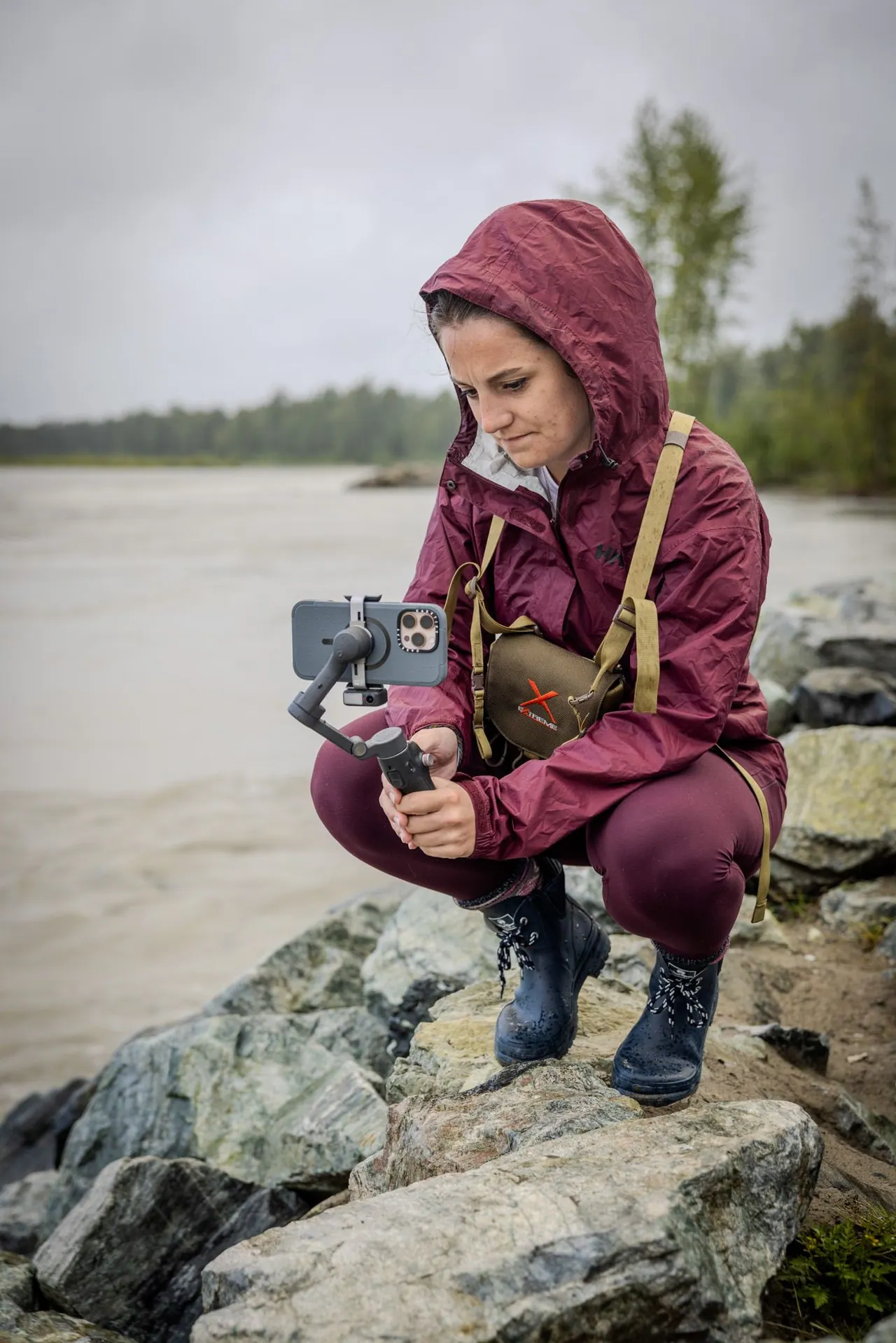 Videographer in a burgundy rain jacket operating a phone gimbal beside a river