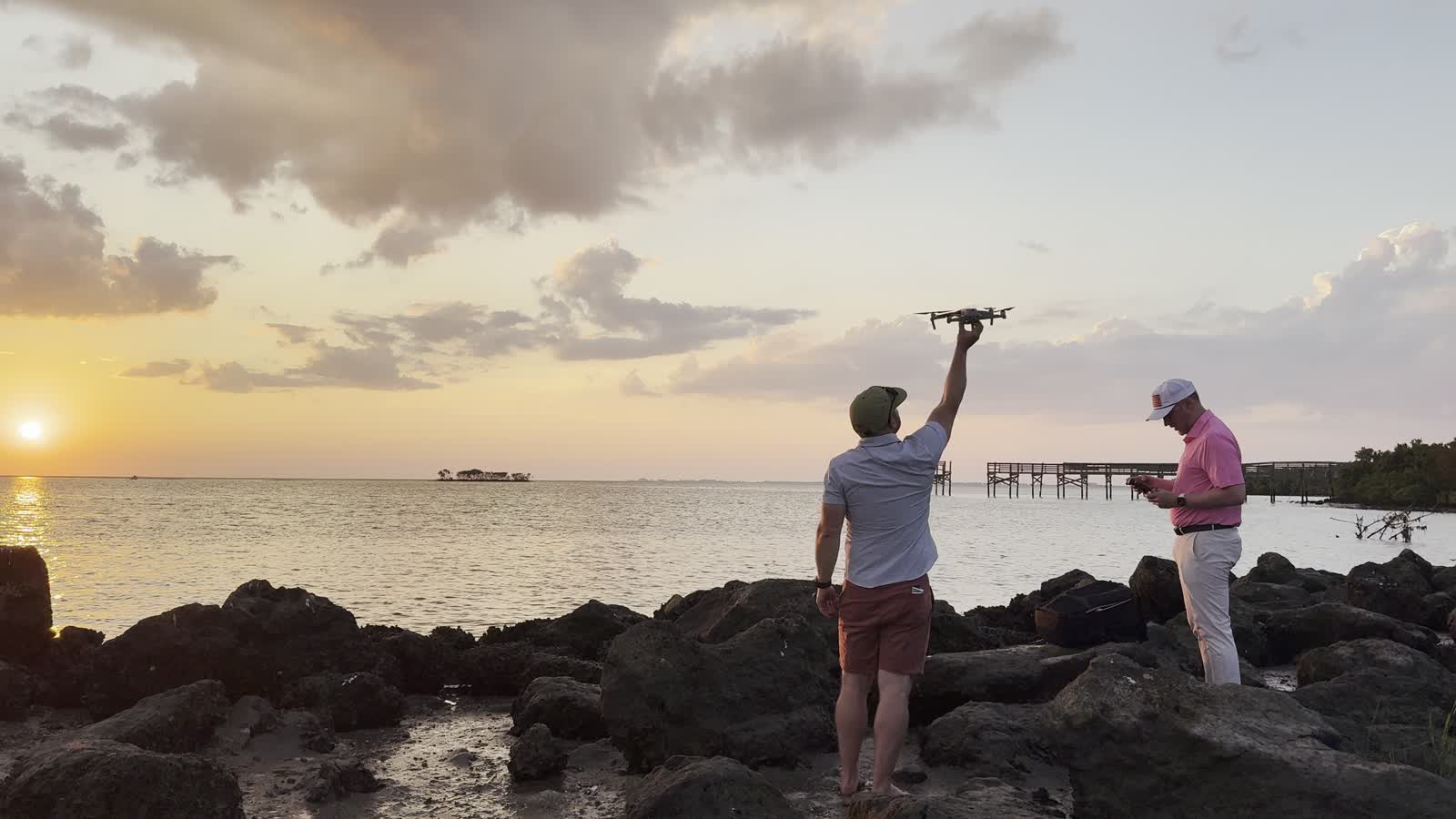 Two people launching a camera drone from a rocky shoreline at sunset over the Gulf.