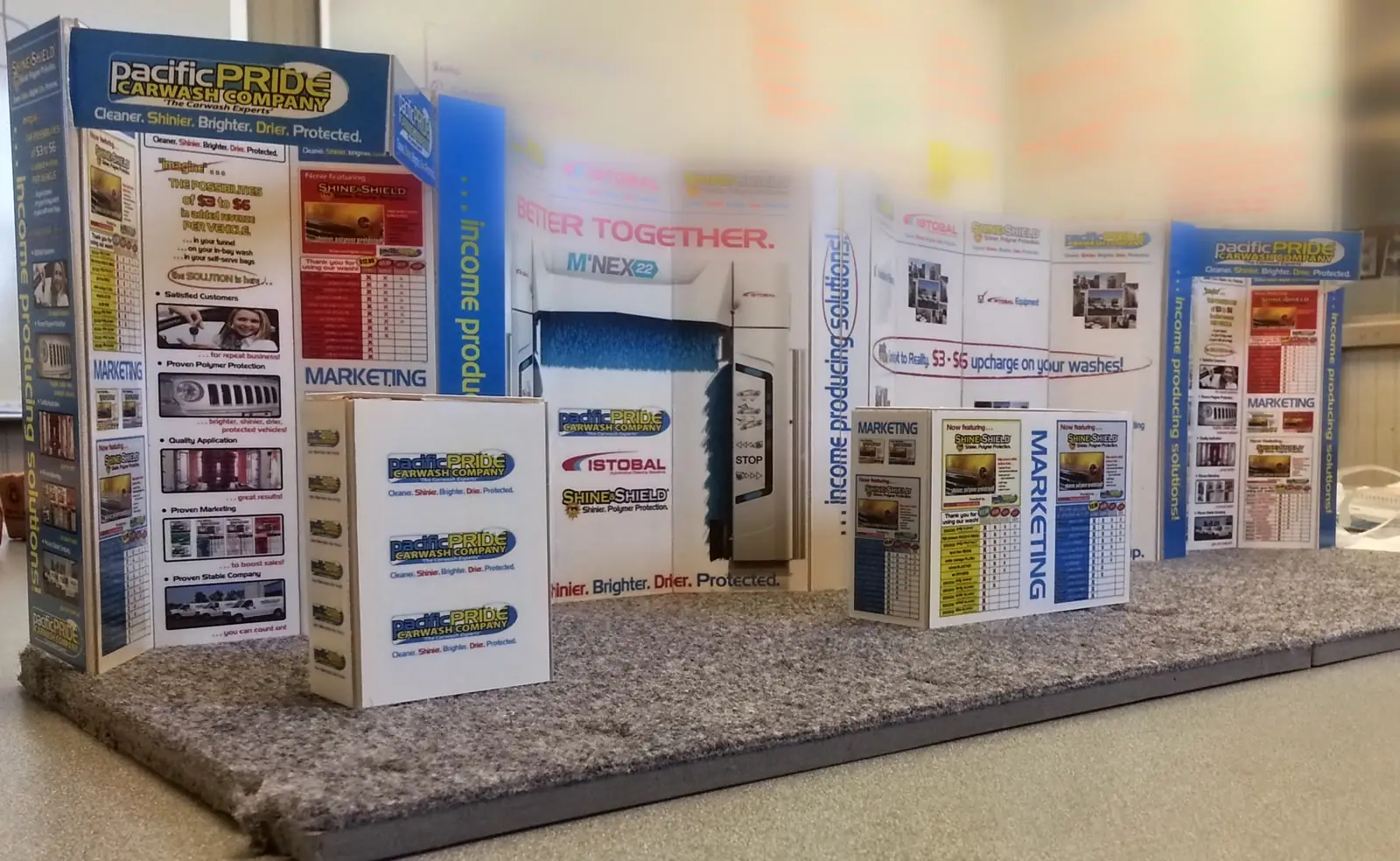 A scale model of a trade show stand on a work table: printed panels standing on a carpeted platform, carrying a carwash company's branding across a run of connected walls